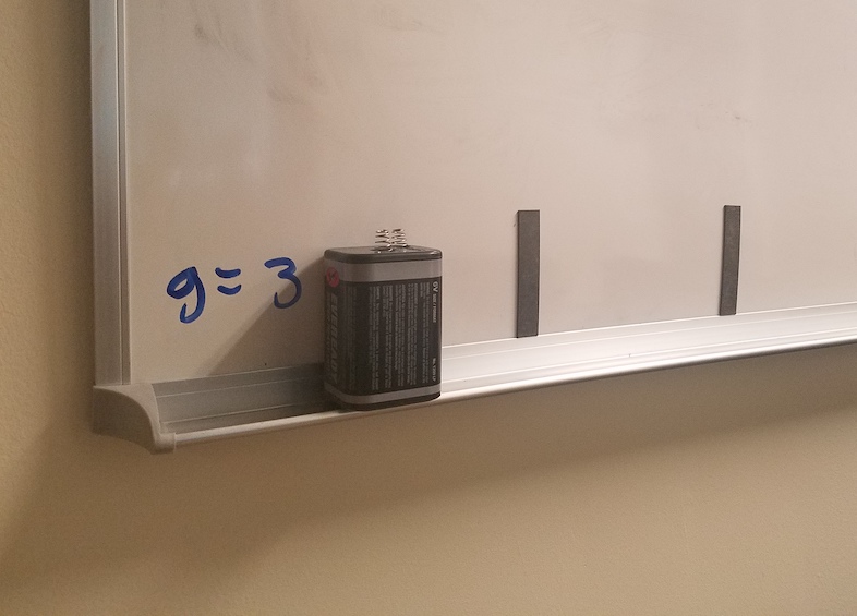 g = 3' written on the bottom corner a whiteboard,
            partially obscured by a battery.