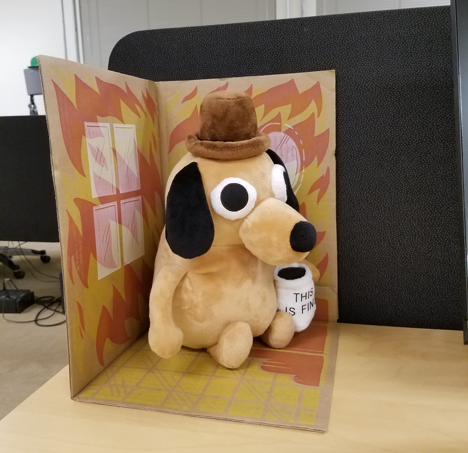 Stuffed dog holding a mug that says 'This is Fine', in a cardboard house with flame pattern