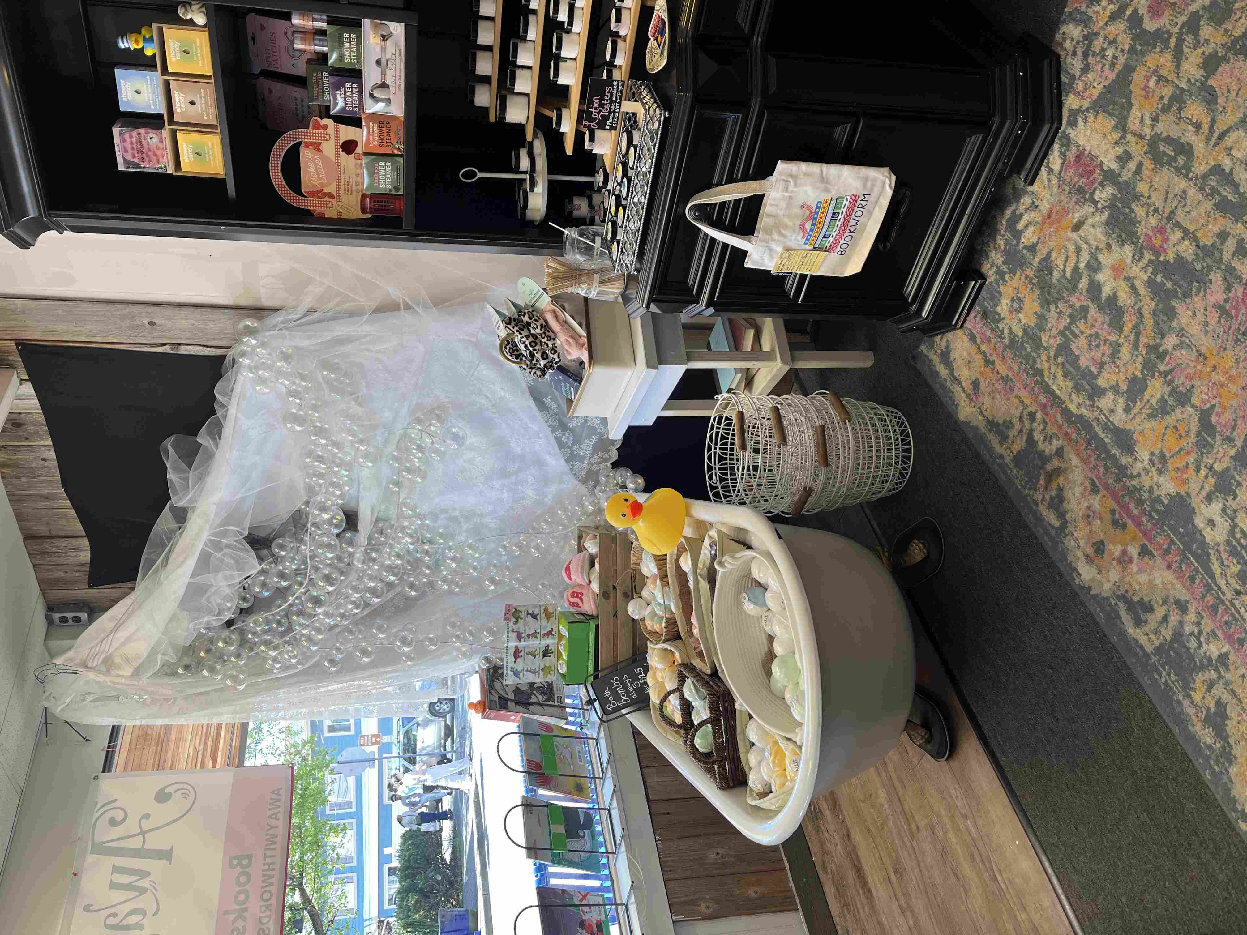 A bathtub filled with bath bombs and a big rubber duck, with bubble decorations draped over it, next to a dark wood shelf full of lotions and shower steamers