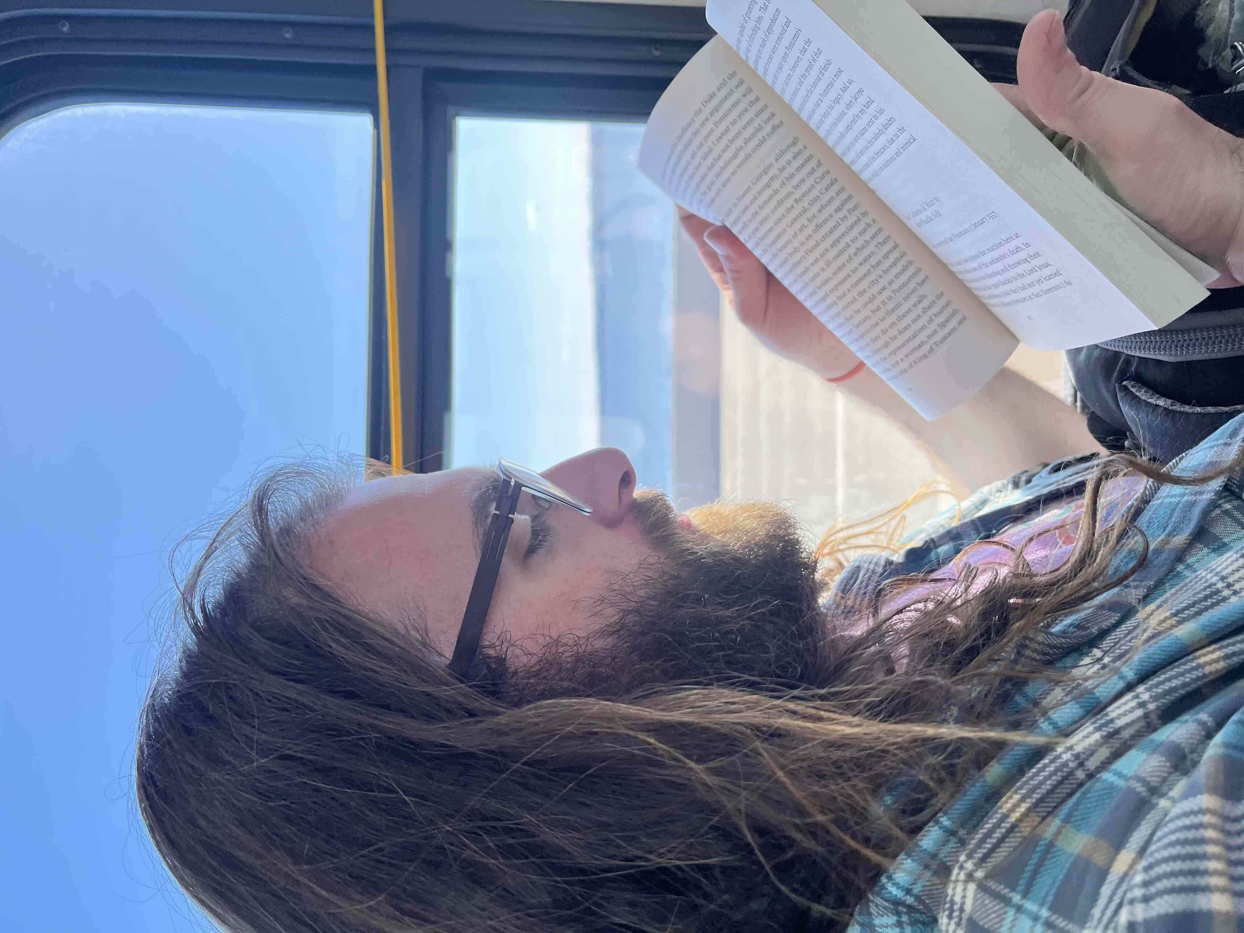 Close up of a bearded man with long hair and glasses reading in front of a bus window, with highway and water in the background.