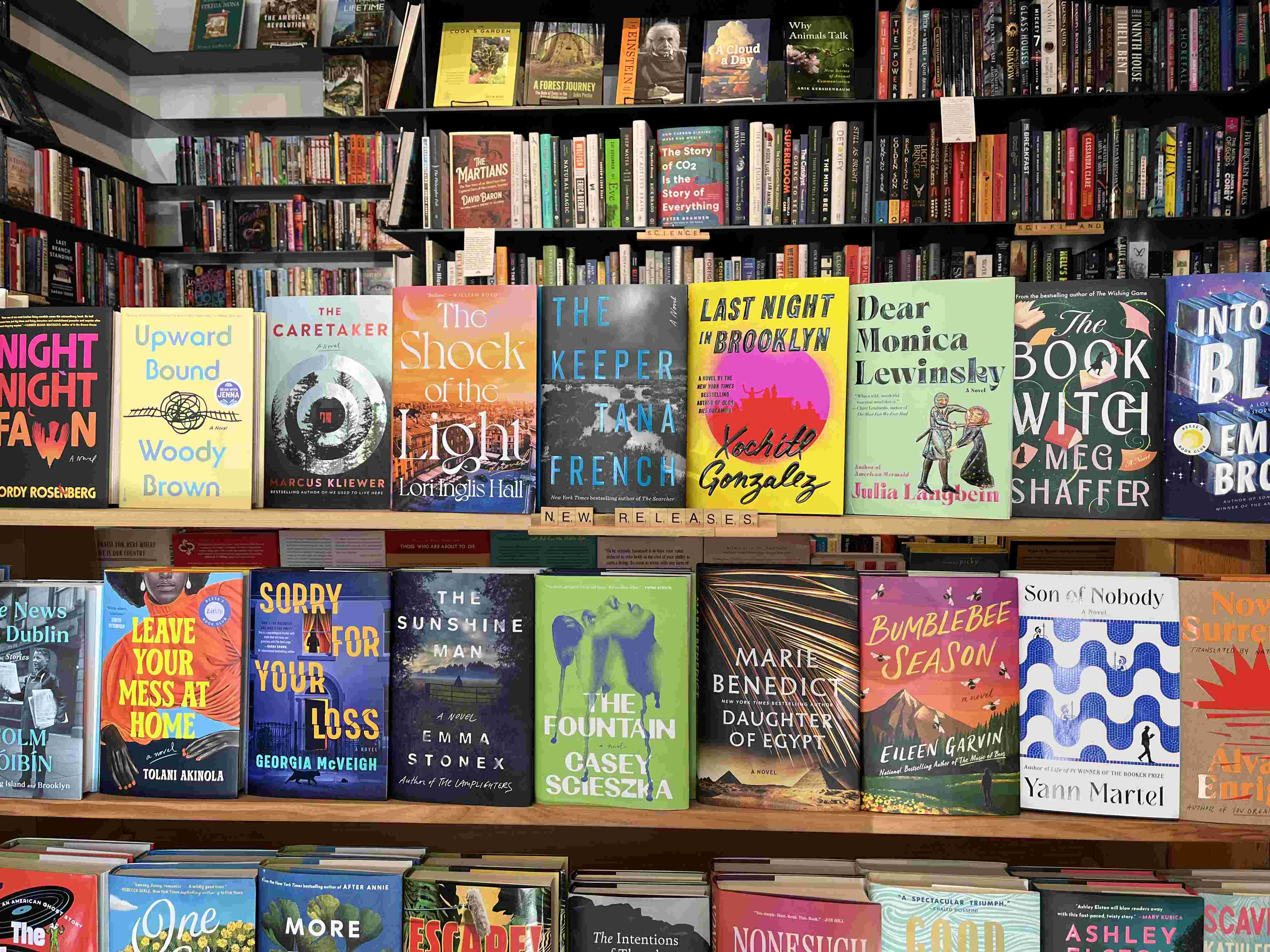 Three shelves filled with colorful books, covers facing out. One shelf has a 'New Releases' label made of Scrabble tiles. More shelves in the background. Some titles: Upward Bound, Last Night in Brooklyn, Dear Monica Lewinsky