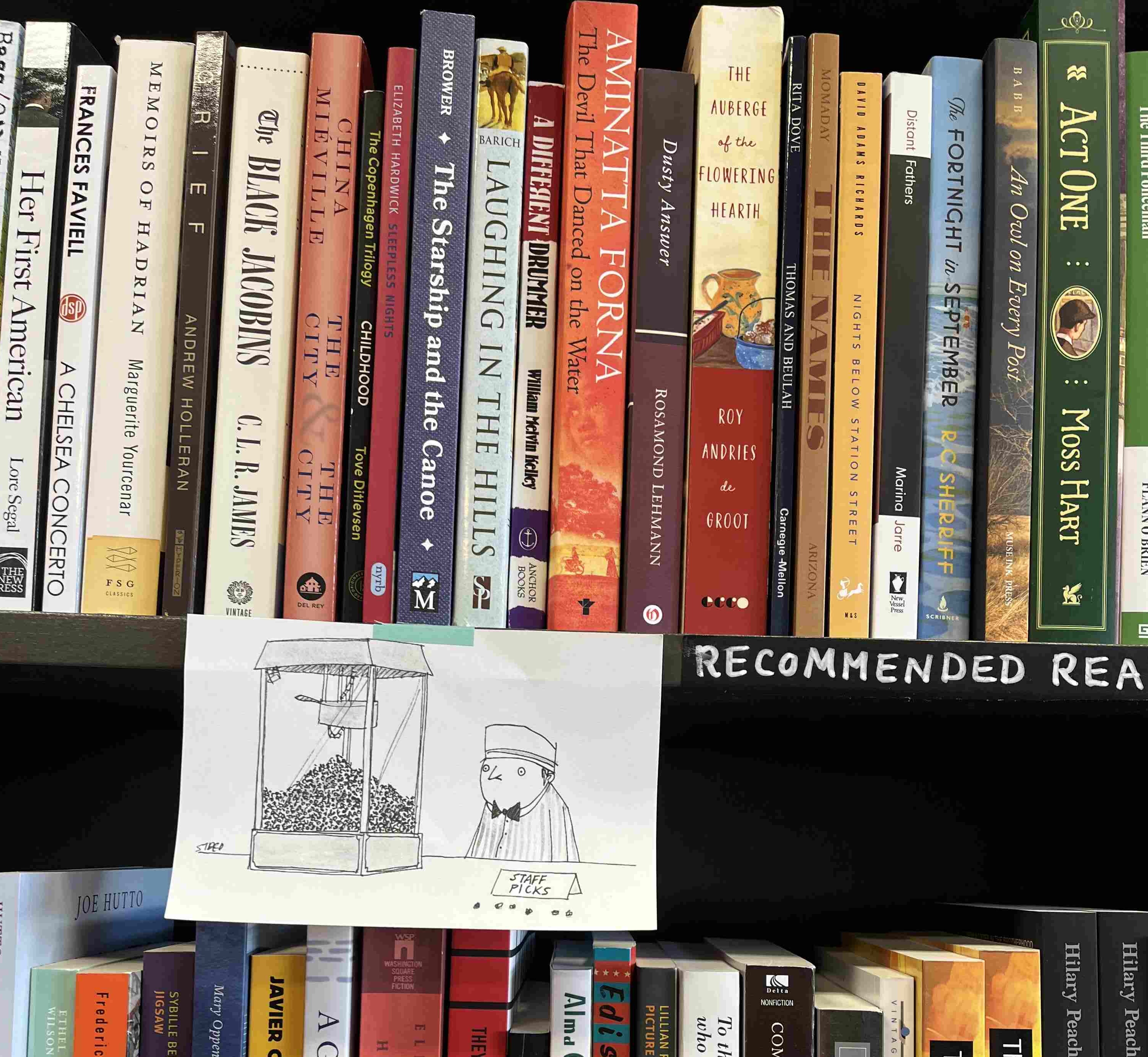 A bookshelf, full of colorful books, including Memoirs of Hadrian, The City & The City, Thomas and Beulah. Taped to the bottom of the shelf, a pen-and-ink drawing taped to it of a man dressed like a movie theater attendant with a dazed expression standing behind a popcorn machine. In the drawing, a sign in front of the man says 'Staff Picks'. Next to the drawing the shelf is labeled 'Recommended Reads'.