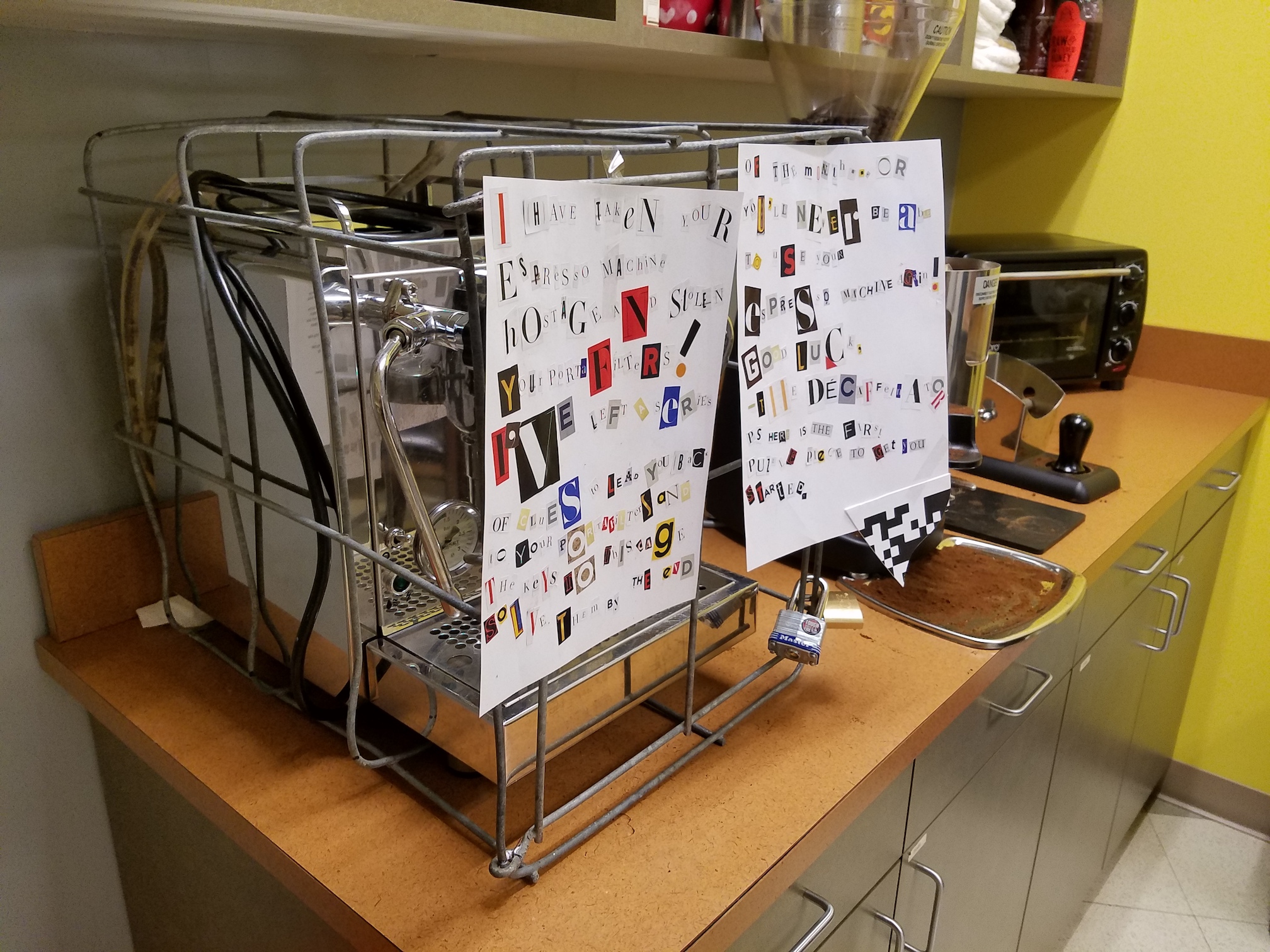 Espresso machine in a metal cage with multiple padlocks. A ransom note made of magazine clippings, and a fragment of a QR code, are taped to the front.